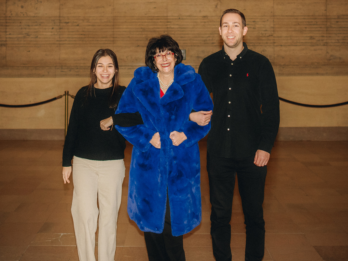 A young woman, her mother in a blue furry coat, and a young man stand for a photo in a train station